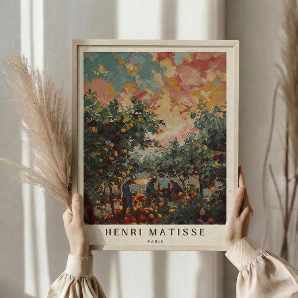 May include: Hands holding a framed art print featuring an impressionist landscape painting with vibrant brushstrokes in blues, oranges, pinks, and greens depicting trees and figures beneath a colorful sky. The print has "Henri Matisse Paris" text below the artwork in a light wooden frame. Dried pampas grass and neutral curtains create a soft, natural backdrop.