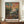 May include: Framed art print featuring a colorful landscape painting with vibrant orange and pink sky, green foliage and fruit trees in foreground, displayed on wooden sideboard next to white ceramic vase and books in elegant room with cream walls and white molding.