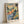 May include: Watercolor landscape print in light wood frame leaning against white wall. Artwork shows Mediterranean hillside village with warm buildings atop colorful terrain in oranges, blues, and greens. Dark cypress trees punctuate the composition. Loose, impressionistic brushwork with flowing washes of color. Artist signature visible in bottom right corner.