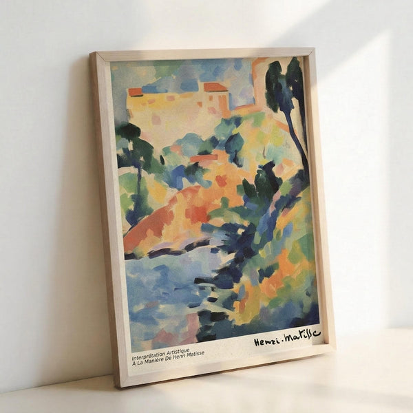 May include: Watercolor landscape print in light wood frame leaning against white wall. Artwork shows Mediterranean hillside village with warm buildings atop colorful terrain in oranges, blues, and greens. Dark cypress trees punctuate the composition. Loose, impressionistic brushwork with flowing washes of color. Artist signature visible in bottom right corner.