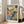 May include: Framed watercolor landscape print in light wood frame leaning against white wall next to beige sofa. Artwork features vibrant blues, greens, oranges and yellows depicting Mediterranean hillside village with cypress trees, rolling terrain and buildings. Fauve-style painting with loose brushstrokes and bold color blocks. Print has white border with text at bottom. Natural lighting from nearby windows creates soft shadows on hardwood floor.