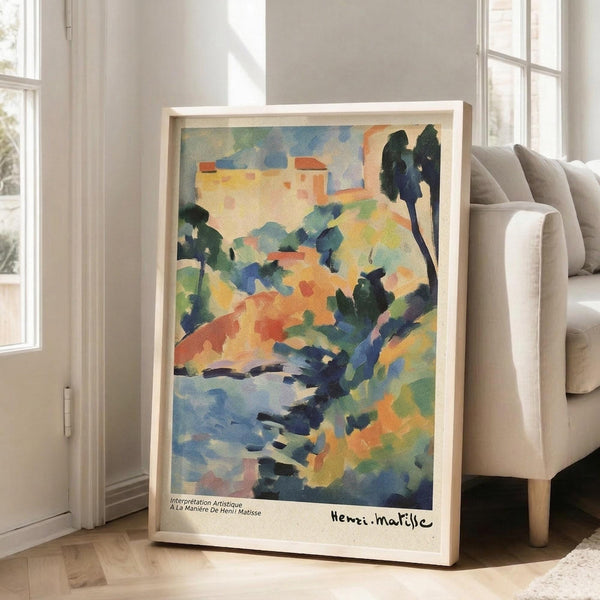 May include: Framed watercolor landscape print in light wood frame leaning against white wall next to beige sofa. Artwork features vibrant blues, greens, oranges and yellows depicting Mediterranean hillside village with cypress trees, rolling terrain and buildings. Fauve-style painting with loose brushstrokes and bold color blocks. Print has white border with text at bottom. Natural lighting from nearby windows creates soft shadows on hardwood floor.