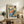 May include: Colorful abstract landscape poster print in light wood frame displayed on rustic wooden sideboard with drawers. Artwork features vibrant blues, oranges, greens, and yellows depicting buildings and trees in Fauvist style. Scene includes cream upholstered chair, potted fern, ceramic vase, and stacked books against white wall in modern living room setting.
