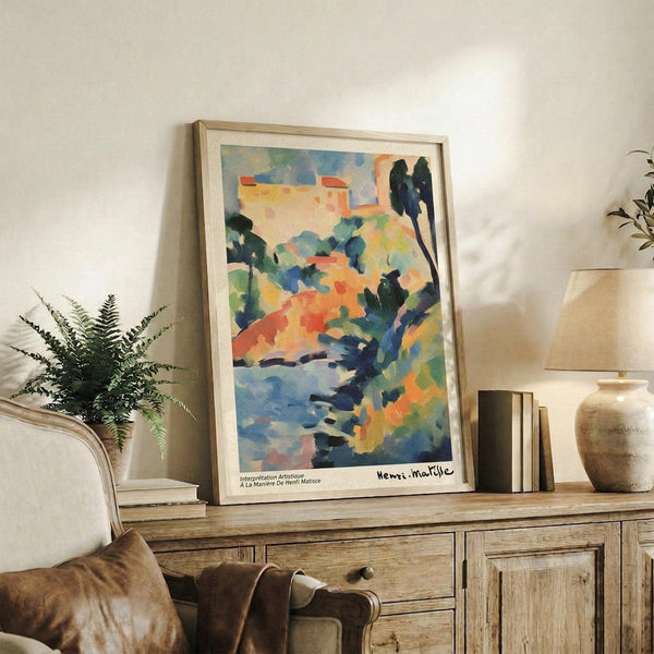 May include: Colorful abstract landscape poster print in light wood frame displayed on rustic wooden sideboard with drawers. Artwork features vibrant blues, oranges, greens, and yellows depicting buildings and trees in Fauvist style. Scene includes cream upholstered chair, potted fern, ceramic vase, and stacked books against white wall in modern living room setting.