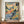May include: A framed colorful abstract landscape poster in Fauve style featuring vibrant blues, oranges, yellows and greens with simplified trees and buildings, displayed on a wooden sideboard next to a white ceramic vase and stack of books in an elegant room with white paneled walls.