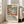May include: Framed art poster featuring a vibrant tropical landscape with palm trees, foliage, and water in bold blues, greens, yellows, and coral tones. The print has exhibition-style text below the colorful artwork. Light wood frame leans against white wall next to beige upholstered sofa in bright, airy room with large windows and natural light streaming across hardwood floors.