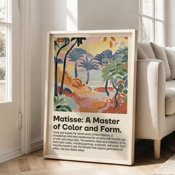 May include: Framed art poster featuring a vibrant tropical landscape with palm trees, foliage, and water in bold blues, greens, yellows, and coral tones. The print has exhibition-style text below the colorful artwork. Light wood frame leans against white wall next to beige upholstered sofa in bright, airy room with large windows and natural light streaming across hardwood floors.