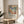 May include: Framed poster print featuring a colorful tropical landscape with palm trees in Matisse's distinctive style, displayed on a round wooden side table next to a beige upholstered chair. Natural sunlight streams across white walls, creating soft shadows. A plant's leaves are visible in the corner, adding to the serene interior setting.