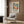 May include: Framed colorful tropical landscape poster with palm trees, vibrant blues, greens, oranges and yellows in Matisse's style, displayed on wood-paneled wall above beige upholstered armchair with geometric patterned pillow, beside brass floor lamp and dark round side table with open book