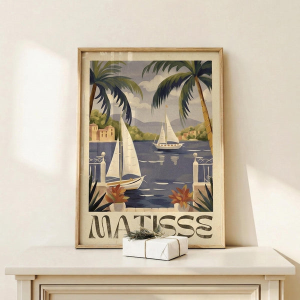 May include: Vintage-style poster print of Mediterranean coastal scene with palm trees, white sailboats on blue water, terracotta buildings, and decorative balcony with potted plants. Text reads "MATISSE" at bottom. Displayed in thin gold frame on light wood surface with small wrapped gift box beside it against cream wall.