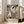 May include: Vintage-style travel poster featuring tropical Mediterranean scene with palm trees, sailboats on blue water, terracotta buildings, and decorative balcony railing in foreground. Poster displays in light wood frame, leaning against beige sofa in bright living room with white walls and large windows. Warm color palette of blues, greens, and earth tones in classic mid-century illustration style.