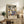May include: Framed vintage-style travel poster featuring tropical coastal scene with palm trees, sailboats on blue water, and "MATISSE" text at bottom, displayed on rustic wooden sideboard alongside fern plant, books, ceramic vase, and cream lamp in neutral-toned living room setting
