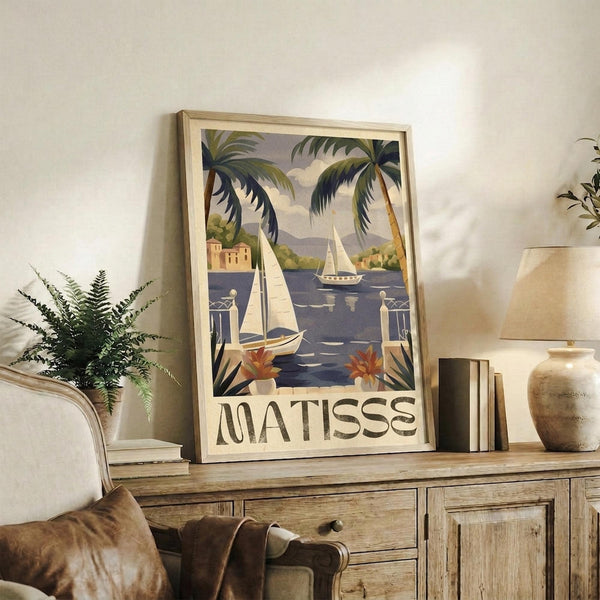 May include: Framed vintage-style travel poster featuring tropical coastal scene with palm trees, sailboats on blue water, and "MATISSE" text at bottom, displayed on rustic wooden sideboard alongside fern plant, books, ceramic vase, and cream lamp in neutral-toned living room setting