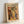 May include: Framed art print in light wood frame leaning against white wall. Poster features "MATISSE" text above colorful still life with various vases, plants, and fruits in Matisse's distinctive cut-out style. Artwork shows warm orange background with yellow, green, black, and blue vessels containing leafy plants, plus scattered circular fruits below. Print has cream-colored background with vibrant, flat color blocks typical of mid-century modern aesthetic.