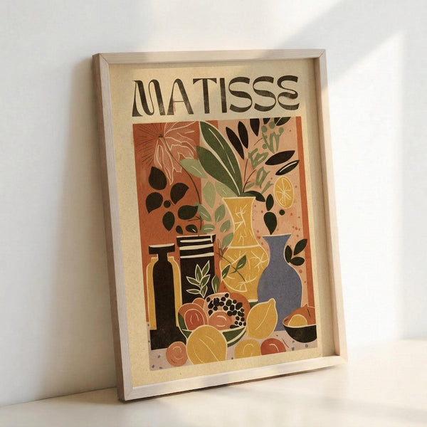 May include: Framed art print in light wood frame leaning against white wall. Poster features "MATISSE" text above colorful still life with various vases, plants, and fruits in Matisse's distinctive cut-out style. Artwork shows warm orange background with yellow, green, black, and blue vessels containing leafy plants, plus scattered circular fruits below. Print has cream-colored background with vibrant, flat color blocks typical of mid-century modern aesthetic.