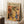 May include: Framed art print in light wood frame leaning against white wall corner. Poster features colorful still life with botanical elements, vases, bottles, and fruits in earthy tones of orange, yellow, green, and blue. Scene includes coffee cup on wooden block and textured beige throw blanket, creating cozy room setting with natural lighting.