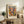 May include: Framed Henri Matisse exhibition poster with colorful abstract cut-out design in blues, oranges, greens, and pinks, displayed on rustic wooden credenza beside fern plant, ceramic vase, and stacked books in neutral-toned living room setting with beige armchair and soft natural lighting.