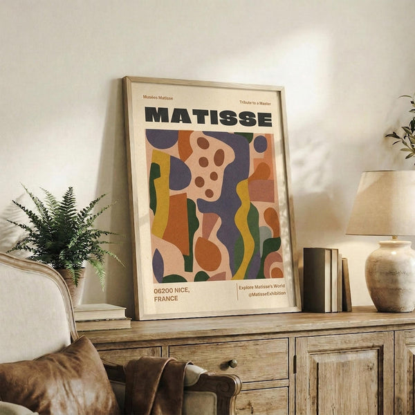May include: Framed Henri Matisse exhibition poster with colorful abstract cut-out design in blues, oranges, greens, and pinks, displayed on rustic wooden credenza beside fern plant, ceramic vase, and stacked books in neutral-toned living room setting with beige armchair and soft natural lighting.