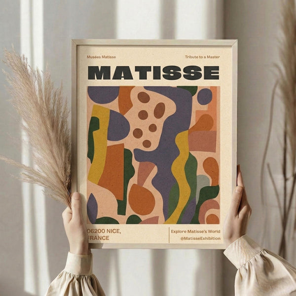 May include: Person in beige ruffled sleeves holding framed Matisse exhibition poster with abstract colorful cut-out shapes in blues, oranges, yellows, and greens. Dried pampas grass stems visible on left side. Light wooden frame against neutral curtained background in soft natural lighting.
