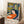 May include: Framed art print leaning against white wall in cozy room setting, showing woman in orange dress with long dark hair surrounded by tropical foliage in blues, yellows, and greens. Matisse-style painting with bold colors and flowing forms. Light wood frame, white mat, positioned near small wooden table with coffee cup and textured rug.