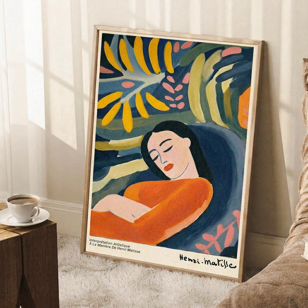 May include: Framed art print leaning against white wall in cozy room setting, showing woman in orange dress with long dark hair surrounded by tropical foliage in blues, yellows, and greens. Matisse-style painting with bold colors and flowing forms. Light wood frame, white mat, positioned near small wooden table with coffee cup and textured rug.