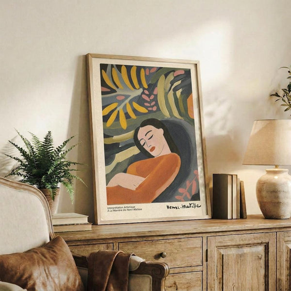 May include: Framed art print displaying a woman with long dark hair in orange clothing against tropical foliage background with yellow and green palm leaves. Print sits on rustic wooden console table alongside potted fern, ceramic vase, stacked books, and table lamp. Cream-colored chair with leather cushion visible in foreground of warmly lit living room setting.