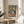 May include: Framed Matisse-inspired art print displayed on round wooden side table beside beige upholstered chair. Artwork features woman with long dark hair in orange top surrounded by tropical foliage in yellow, green, and coral tones. Natural lighting creates soft shadows across neutral-toned room with plant leaves visible in corner.