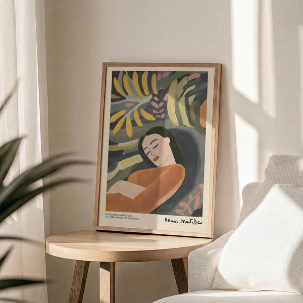 May include: Framed Matisse-inspired art print displayed on round wooden side table beside beige upholstered chair. Artwork features woman with long dark hair in orange top surrounded by tropical foliage in yellow, green, and coral tones. Natural lighting creates soft shadows across neutral-toned room with plant leaves visible in corner.