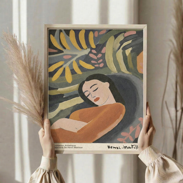 May include: Hands holding a framed art print featuring a reclining woman with dark hair in an orange garment, surrounded by tropical foliage in yellow, green, and coral tones. The print has a light wooden frame and signature at bottom. Person wears cream-colored clothing and holds dried pampas grass. Soft natural lighting creates gentle shadows on neutral background.