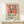 May include: Framed poster featuring a colorful Henri Matisse-style interior scene with red curtains framing a window view of sailboats, potted plants, and decorative elements in vibrant blues, greens, and oranges. Exhibition-style text below describes Matisse as a master of color and form. Natural wood frame displayed on light wood shelf next to small wrapped gift with greenery accent against cream wall.