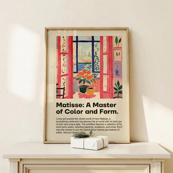 May include: Framed poster featuring a colorful Henri Matisse-style interior scene with red curtains framing a window view of sailboats, potted plants, and decorative elements in vibrant blues, greens, and oranges. Exhibition-style text below describes Matisse as a master of color and form. Natural wood frame displayed on light wood shelf next to small wrapped gift with greenery accent against cream wall.
