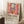 May include: Framed art poster featuring a colorful Matisse-style interior scene with red open window shutters, orange flowers in vases, and view of sailboats on water. Exhibition text reads "Matisse: A Master of Color and Form" below the artwork. Poster leans against white wall next to white coffee cup on wooden surface and beige textured throw blanket in bright, airy room setting.