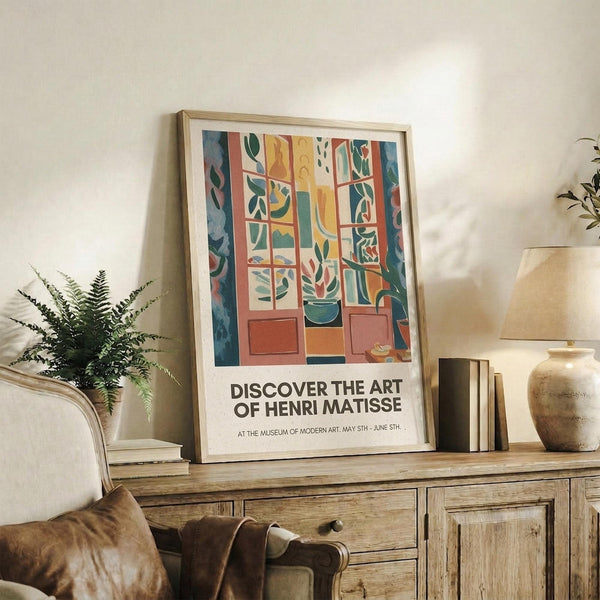 May include: Framed Henri Matisse exhibition poster featuring colorful collage artwork with geometric patterns, botanical motifs, and warm tones of orange, green, and blue. The poster sits on a rustic wooden sideboard alongside books, a ceramic vase, and fern plant, with cream upholstered furniture visible in a neutral-toned living room setting.