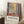 May include: A framed art poster leaning against white walls in a cozy room setting. The artwork shows a colorful window scene with geometric shapes, plants, and vibrant blues, greens, yellows and coral tones. Text at bottom reads "DISCOVER THE ART OF HENRI MATISSE" with museum exhibition details. A coffee cup sits on a wooden block nearby, with soft textured rugs and neutral bedding visible.