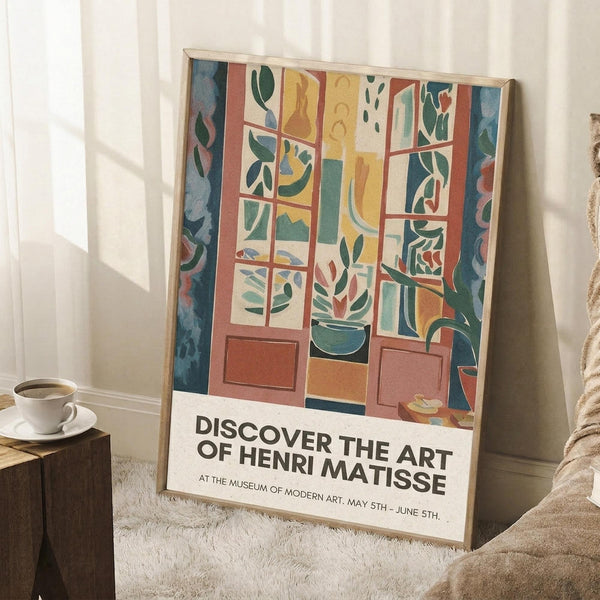 May include: A framed art poster leaning against white walls in a cozy room setting. The artwork shows a colorful window scene with geometric shapes, plants, and vibrant blues, greens, yellows and coral tones. Text at bottom reads "DISCOVER THE ART OF HENRI MATISSE" with museum exhibition details. A coffee cup sits on a wooden block nearby, with soft textured rugs and neutral bedding visible.