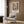 May include: Framed Henri Matisse exhibition poster hanging on wood-paneled wall above beige upholstered armchair with geometric patterned pillow. Poster features colorful abstract interior scene with window, plants, and furniture in oranges, blues, and greens. Modern reading corner with brass floor lamp and dark round side table with open book. Warm lighting and neutral curtains complete the contemporary living space mockup.