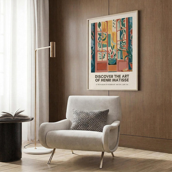 May include: Framed Henri Matisse exhibition poster hanging on wood-paneled wall above beige upholstered armchair with geometric patterned pillow. Poster features colorful abstract interior scene with window, plants, and furniture in oranges, blues, and greens. Modern reading corner with brass floor lamp and dark round side table with open book. Warm lighting and neutral curtains complete the contemporary living space mockup.