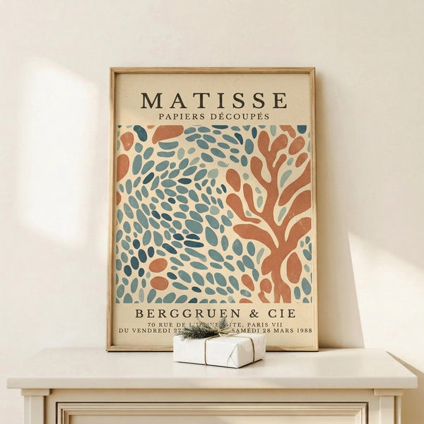 May include: Framed Matisse exhibition poster with abstract coral and teal leaf patterns on cream background, displayed on light wood console table next to small white gift box with greenery, against neutral wall with soft natural lighting.