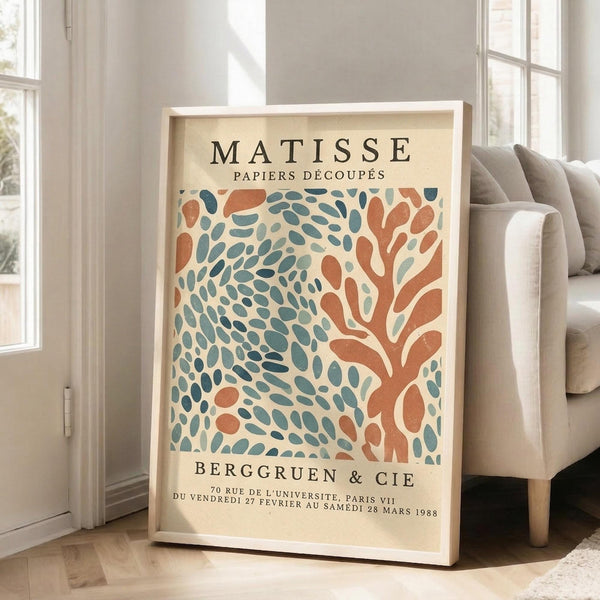 May include: Framed vintage-style exhibition poster featuring abstract coral and teal organic shapes in Matisse's cut-out style, displayed on wooden floor beside beige sofa in bright living room with large windows. Cream-colored frame complements the warm neutral interior.