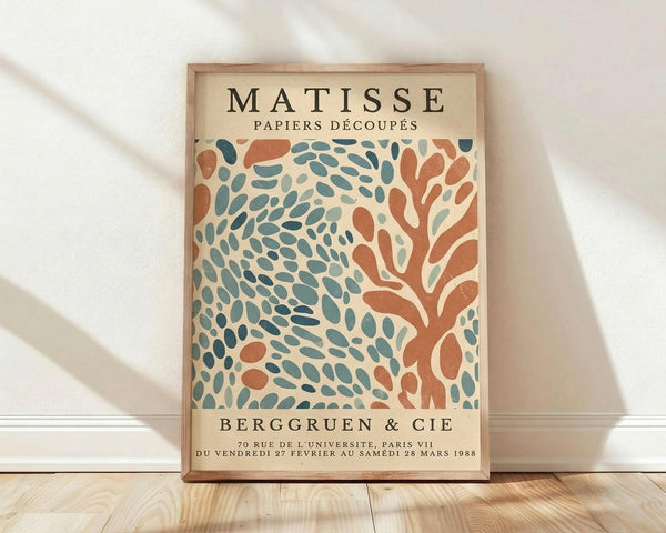 May include: Framed vintage-style exhibition poster featuring abstract organic shapes in coral orange and teal blue on cream background. The artwork shows flowing, leaf-like cutout patterns reminiscent of Matisse's paper collage style. Wooden frame leans against white wall on light wood flooring with soft natural lighting creating gentle shadows.