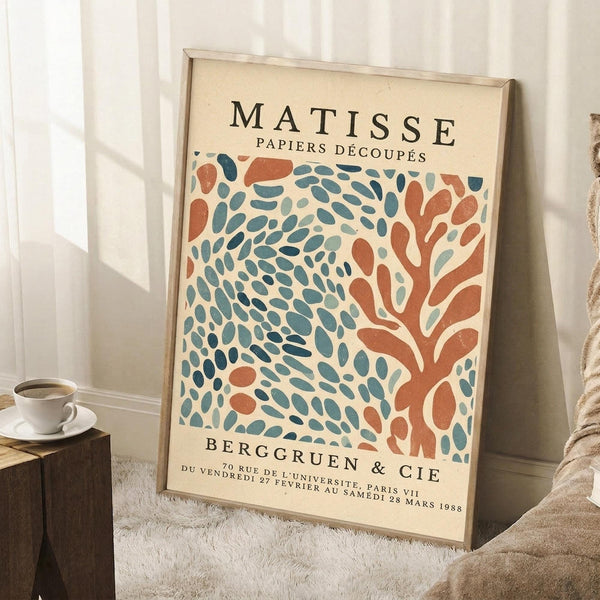 May include: Framed vintage-style art print featuring abstract coral and teal leaf patterns in Matisse's cut-out style, leaning against white wall corner next to wooden side table with white coffee cup and saucer, positioned on textured white rug with beige throw blanket visible at edge.