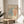 May include: Framed Matisse exhibition poster with abstract coral and teal organic shapes on cream background, displayed on round wooden stool next to beige upholstered chair. Natural sunlight creates soft shadows across white walls. Green plant leaves visible in corner. Warm, minimalist home decor styling with neutral tones throughout the scene.
