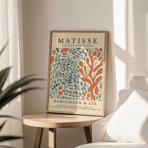 May include: Framed Matisse exhibition poster with abstract coral and teal organic shapes on cream background, displayed on round wooden stool next to beige upholstered chair. Natural sunlight creates soft shadows across white walls. Green plant leaves visible in corner. Warm, minimalist home decor styling with neutral tones throughout the scene.