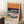 May include: Framed vintage-style poster featuring abstract seascape with layered waves in teal, orange, purple and blue tones against orange sky, displayed leaning against white wall corner next to wooden side table with coffee cup, positioned on textured white rug beside beige blanket in bright natural lighting