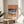 May include: A framed art print featuring an abstract seascape with layered waves in blues, greens, and purples beneath an orange sunset sky, displayed on a light wood round side table next to a beige upholstered chair. Sunlight streams across white walls creating geometric shadows, with a plant's leaves visible in the corner of the bright, modern room setting.