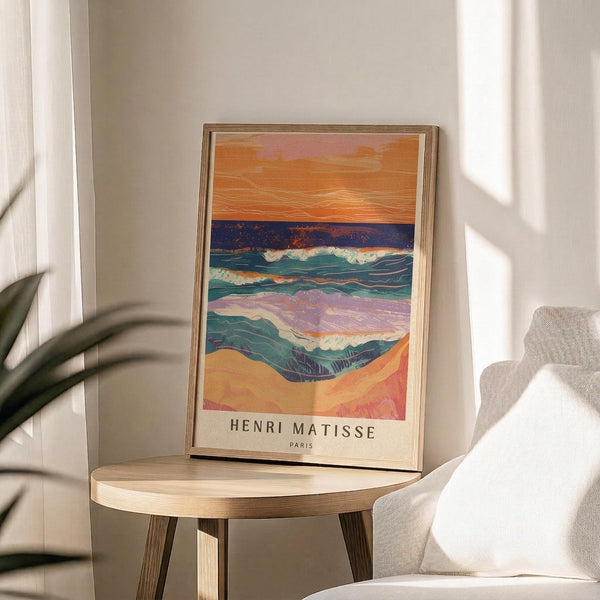 May include: A framed art print featuring an abstract seascape with layered waves in blues, greens, and purples beneath an orange sunset sky, displayed on a light wood round side table next to a beige upholstered chair. Sunlight streams across white walls creating geometric shadows, with a plant's leaves visible in the corner of the bright, modern room setting.