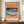 May include: Framed art print displaying an abstract coastal landscape with flowing waves in teal and purple, golden sandy beach, and warm orange-pink sunset sky with flowing linear details. The print features "HENRI MATISSE PARIS" text at bottom. Shown in thin dark wooden frame on walnut credenza beside white ceramic vase and stacked books in neutral-toned room with white paneled walls.