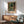 May include: Henri Matisse poster print in wooden frame hanging on white wall above mid-century modern wooden credenza. Artwork depicts colorful tropical garden scene with lush plants, flowers, and architectural elements in vibrant greens, oranges, and reds. Room features sage green upholstered armchair with wooden frame, small round coffee table, and ceramic table lamp, all in warm natural lighting with plant shadows.
