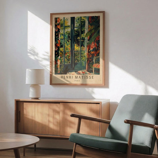 May include: Henri Matisse poster print in wooden frame hanging on white wall above mid-century modern wooden credenza. Artwork depicts colorful tropical garden scene with lush plants, flowers, and architectural elements in vibrant greens, oranges, and reds. Room features sage green upholstered armchair with wooden frame, small round coffee table, and ceramic table lamp, all in warm natural lighting with plant shadows.