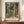 May include: Framed Henri Matisse exhibition poster featuring vibrant tropical scene with French doors opening to lush garden filled with palm fronds, colorful flowers in greens, oranges, and corals. Poster displays on wooden sideboard beside white ceramic vase and stacked books in elegant room with cream walls and white molding.